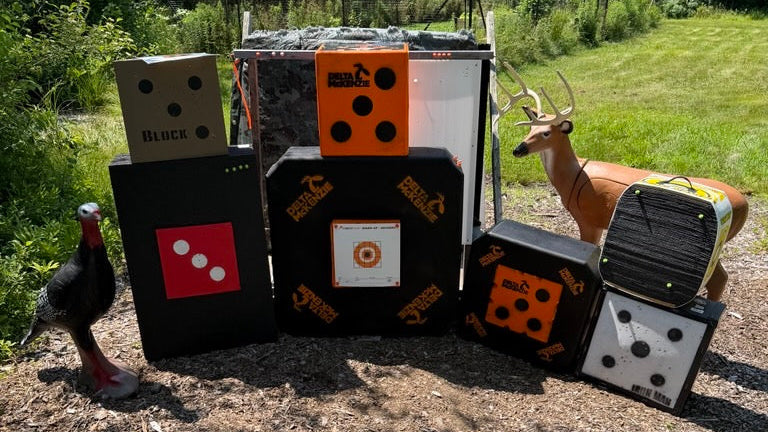 A set of outdoor target practice items including BIGShot and Delta McKenzie black foam blocks, a turkey target, a deer target, arranged in a backyard range on a dirt and mulch ground with trees in the background.